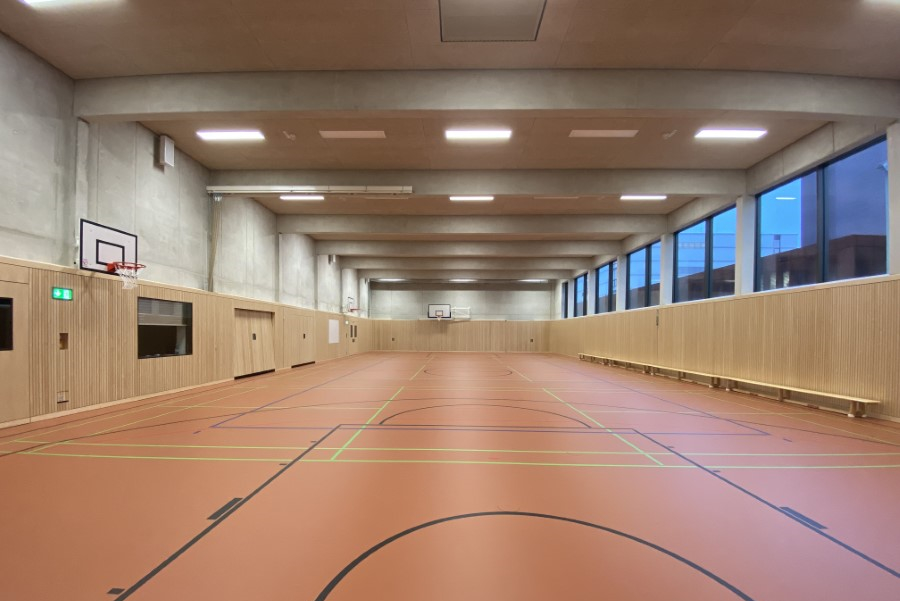 Zu sehen ist die Sporthalle von innen der Boden ist Orange und hat mehrere Abgrenzungslinien während der untere Teil der Wände aus Holzlatten besteht und darüber noch Basketballkörbe und und die Fenster nach außen zu sehen sind.
